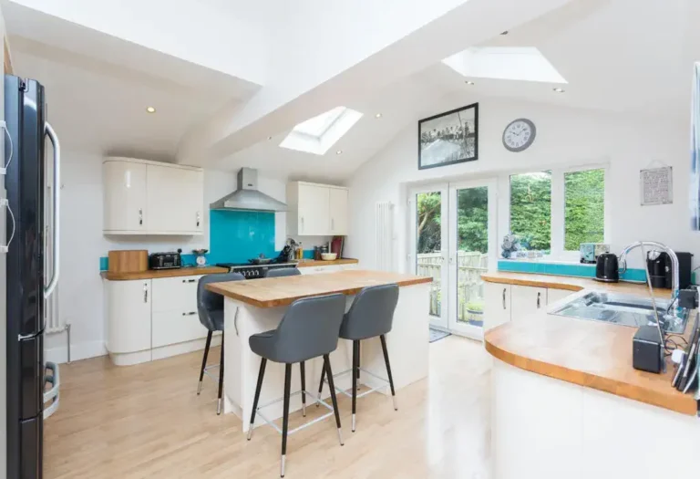 The modern kitchen features skylights, white cabinets, and an island with stools, set against light wood floors. A turquoise backsplash behind the stove adds a pop of color, while a clock and framed picture create charm above the doors—an ideal job template for interior inspiration.